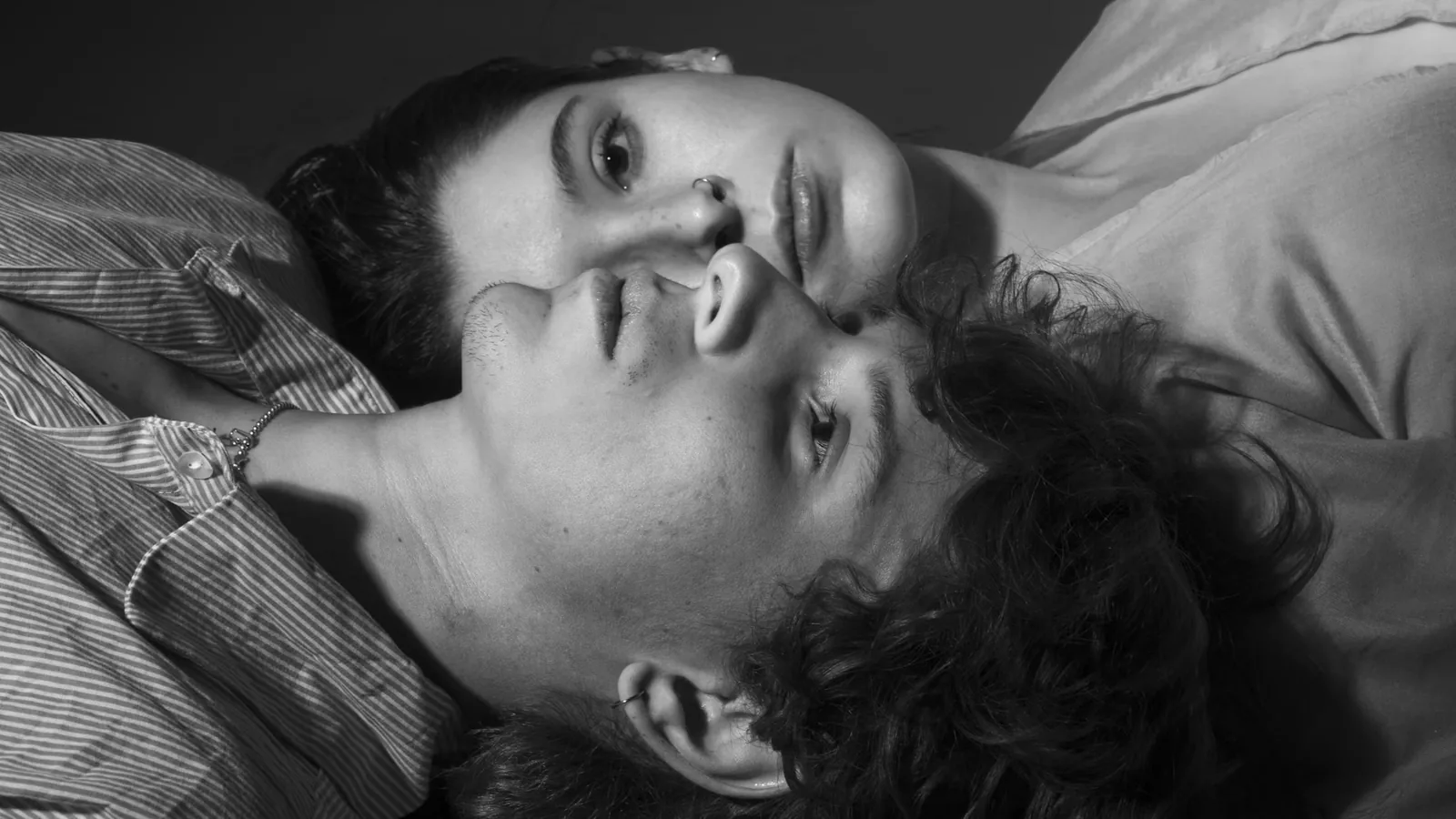 Black and white photograph of a young couple lying side by side with their heads close together, one looking toward the camera