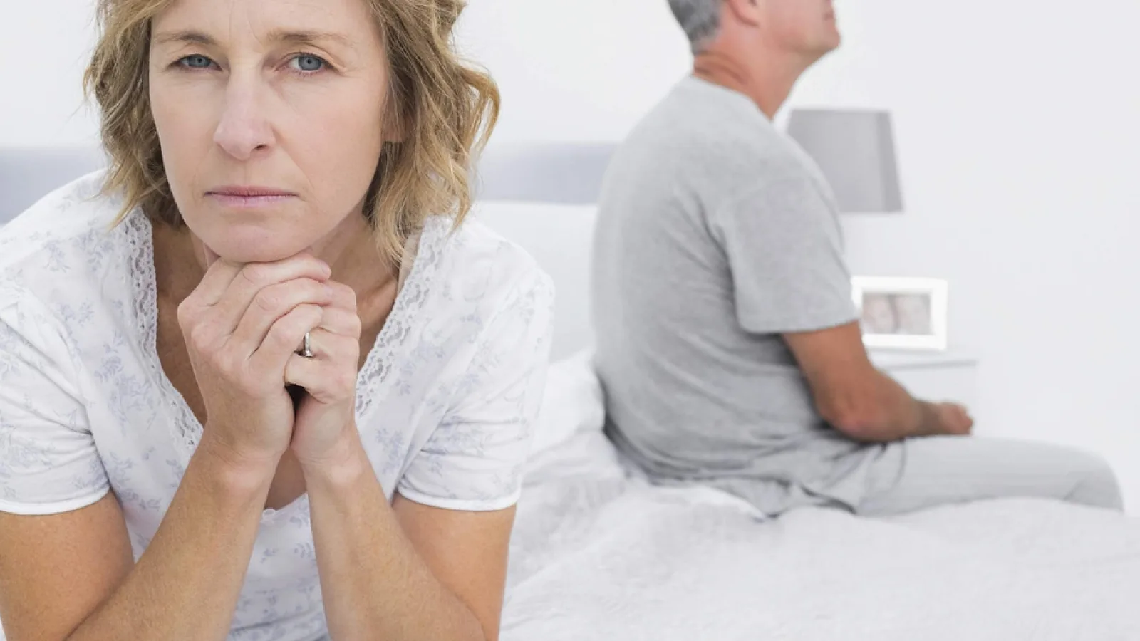 A worried woman resting her chin on her hands while a man sits on the far side of the bed