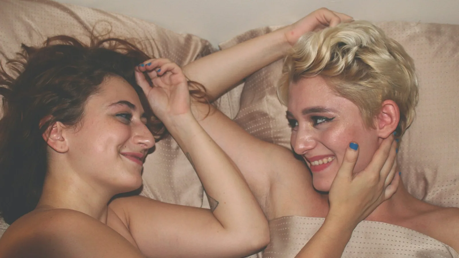 Two women lying in bed laughing together, one touching the other's face