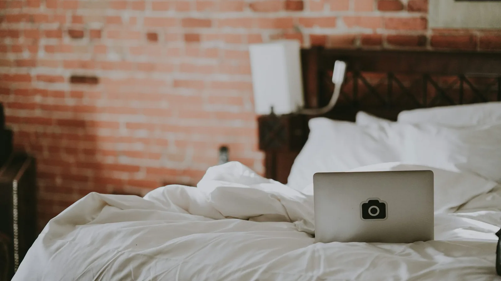 An unmade bed with an open laptop on the duvet, in a loft bedroom with a brick wall