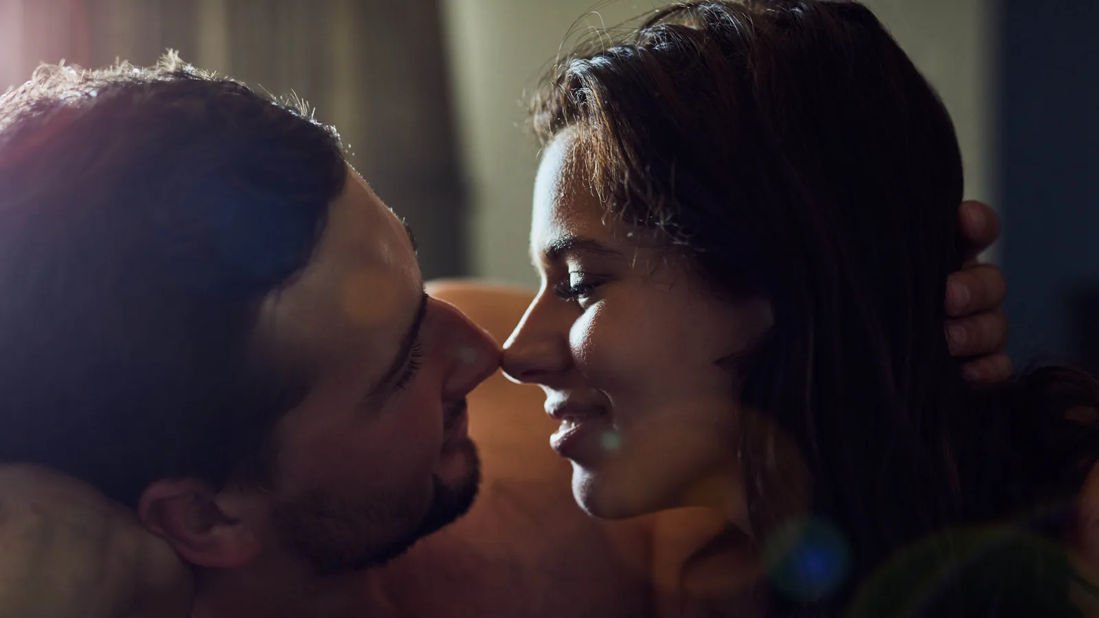 A man and a woman with bare shoulders lying face to face with noses almost touching, his hand cradling her head, in warm low cinematic light