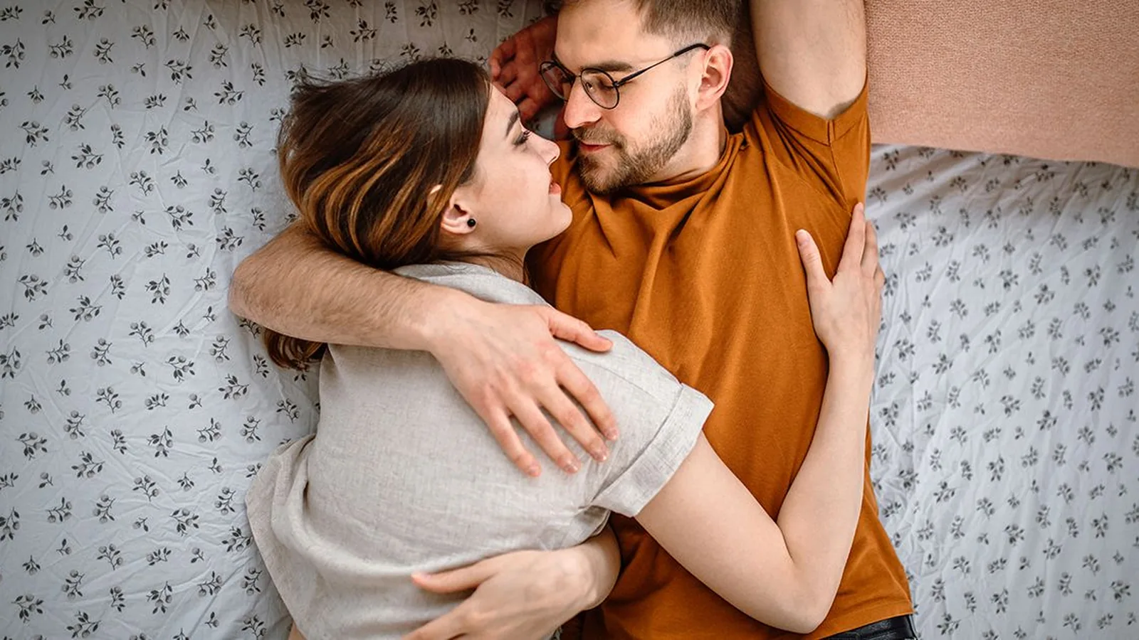 A fully clothed couple lying face to face on a bed with white floral bedding, gazing at each other in an affectionate embrace