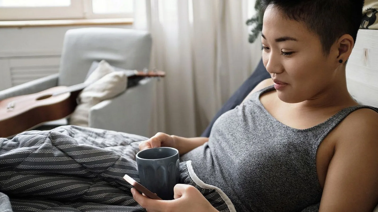 A relaxed woman in a tank top lying in bed at home, holding a smartphone in one hand and a mug in the other, with an armchair and a guitar behind her