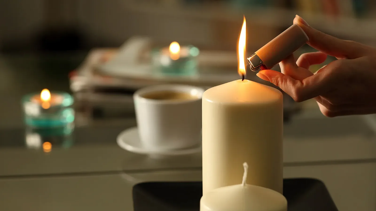 A hand lighting a cream pillar candle beside a cup of coffee
