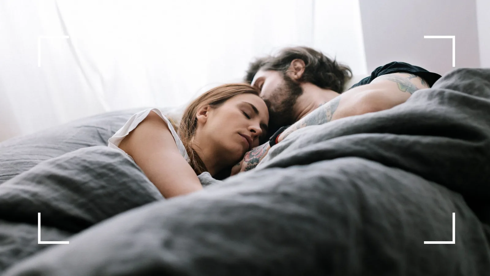 A bearded man lying close behind a woman in a white t-shirt on gray bedding, both resting with eyes closed in soft daylight from sheer curtains