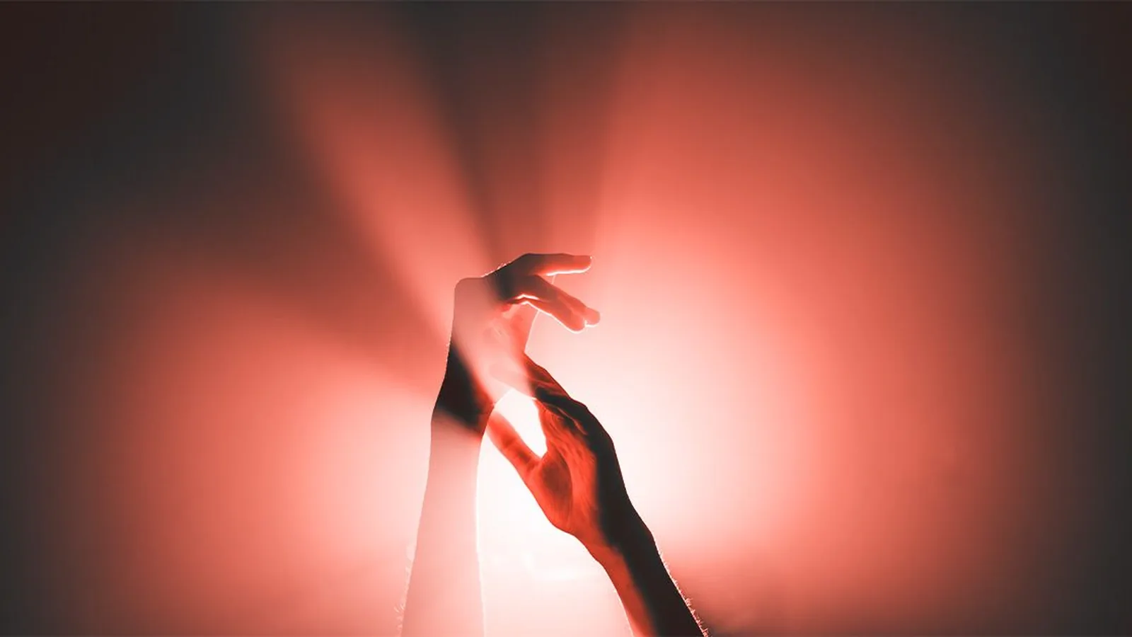 Two hands and forearms reaching toward each other in silhouette against a warm red backlit glow with light rays flaring outward