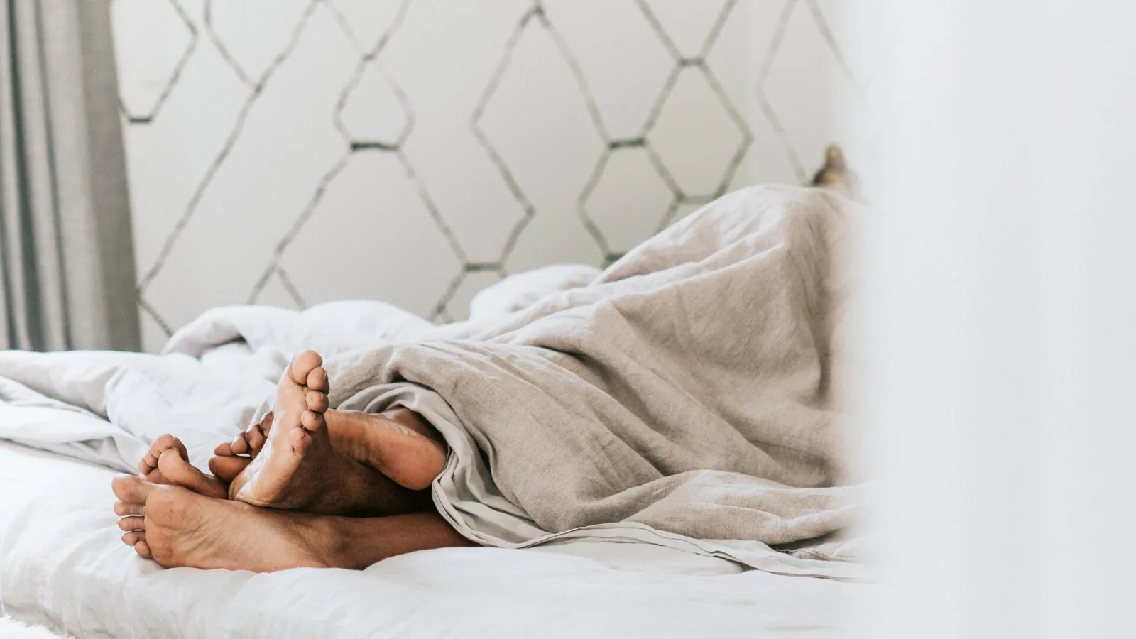 A couple's bare feet poking out from under a beige duvet on a bed against a white wall with a subtle geometric pattern