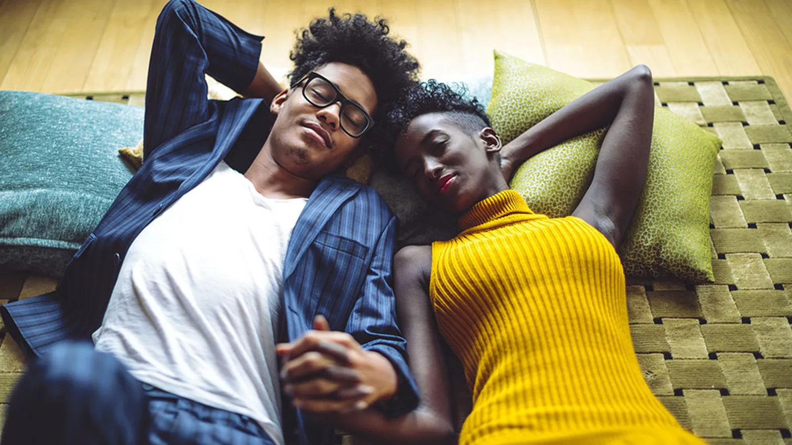 A fully clothed young couple lying head-to-head on pillows on a wooden floor, holding hands with eyes closed